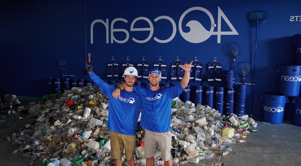 Two men staning in from of heap of garbage in a warehouse with shirts that say 4Ocean and the 4Ocean logo in the background on the wall.